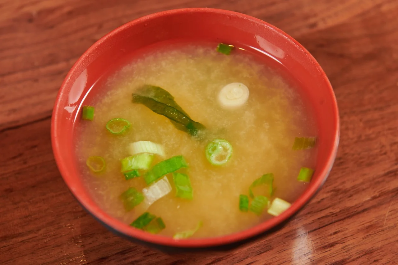 A red lacquer bowl of miso soup with sliced scallion and a strip of wakame, on a wooden table.
