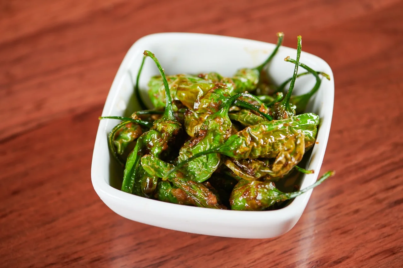 Blistered shishito peppers in a square white bowl, glossed with sauce, on a wooden table.
