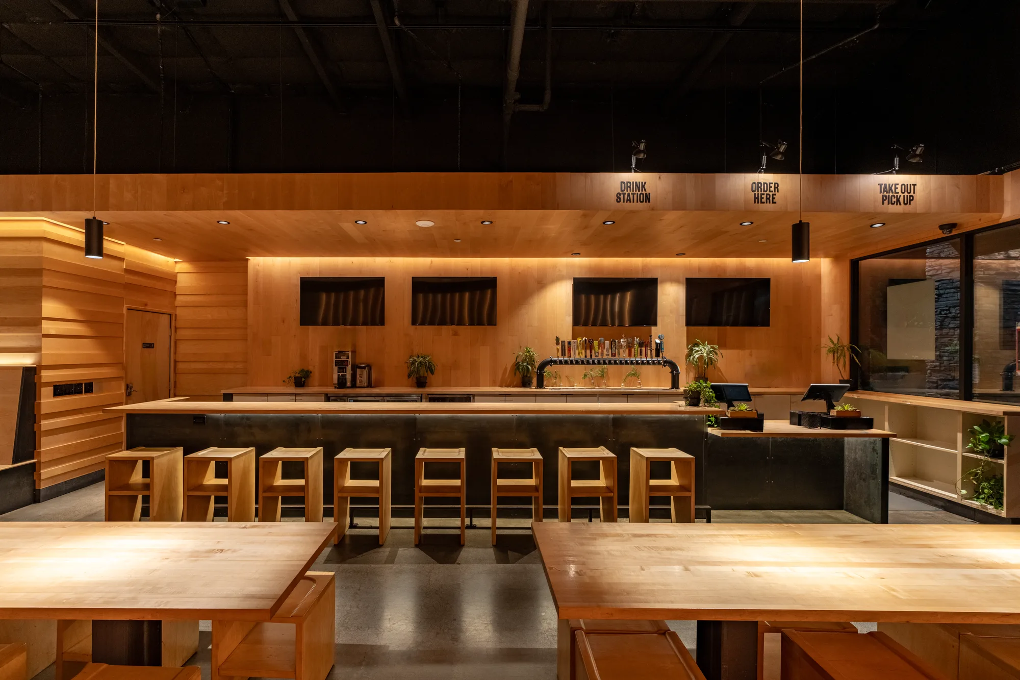 The service counter at College Heights, head on. A row of maple stools stands along a dark counter under a bank of beer taps, with black signs above reading Drink Station, Order Here and Take Out Pick Up, and communal tables in the foreground.