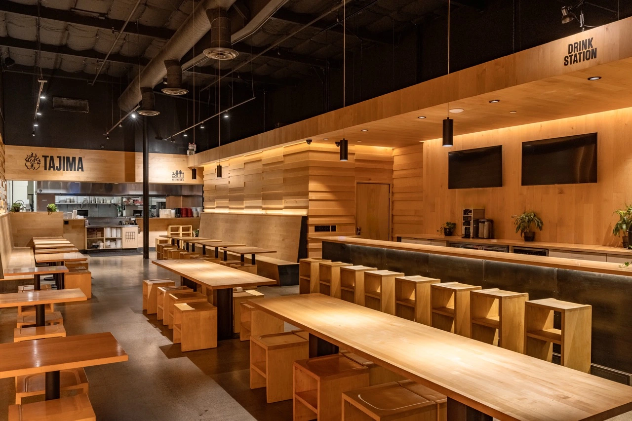 The College Heights dining room before service. Long maple communal tables and box stools run down the room under an exposed black ceiling, with the open kitchen visible past the counter at the far end.
