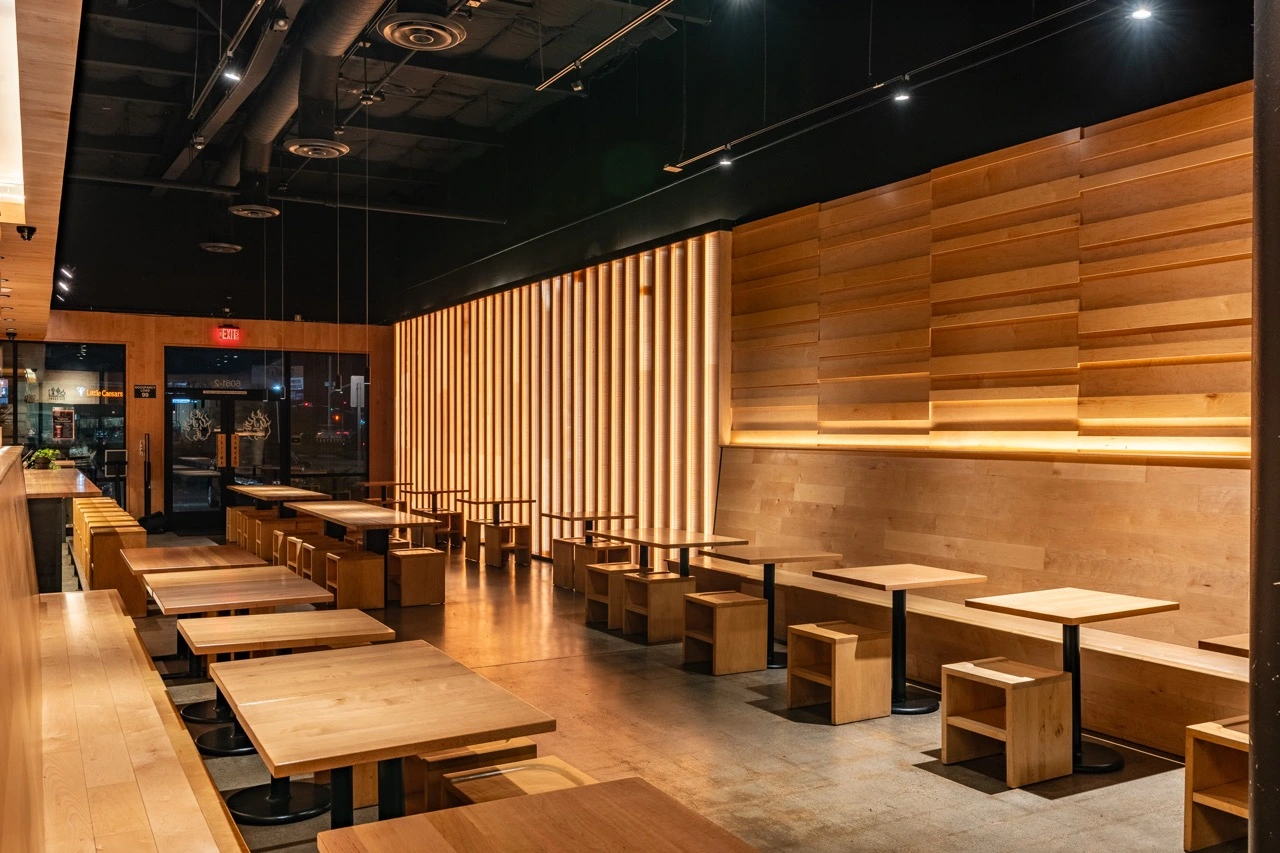 The window side of the College Heights dining room before service. Two-top tables and low maple benches run along the glass toward the front door, with a floor to ceiling screen of vertical maple slats dividing the room and stepped maple shelving lit from behind on the right.