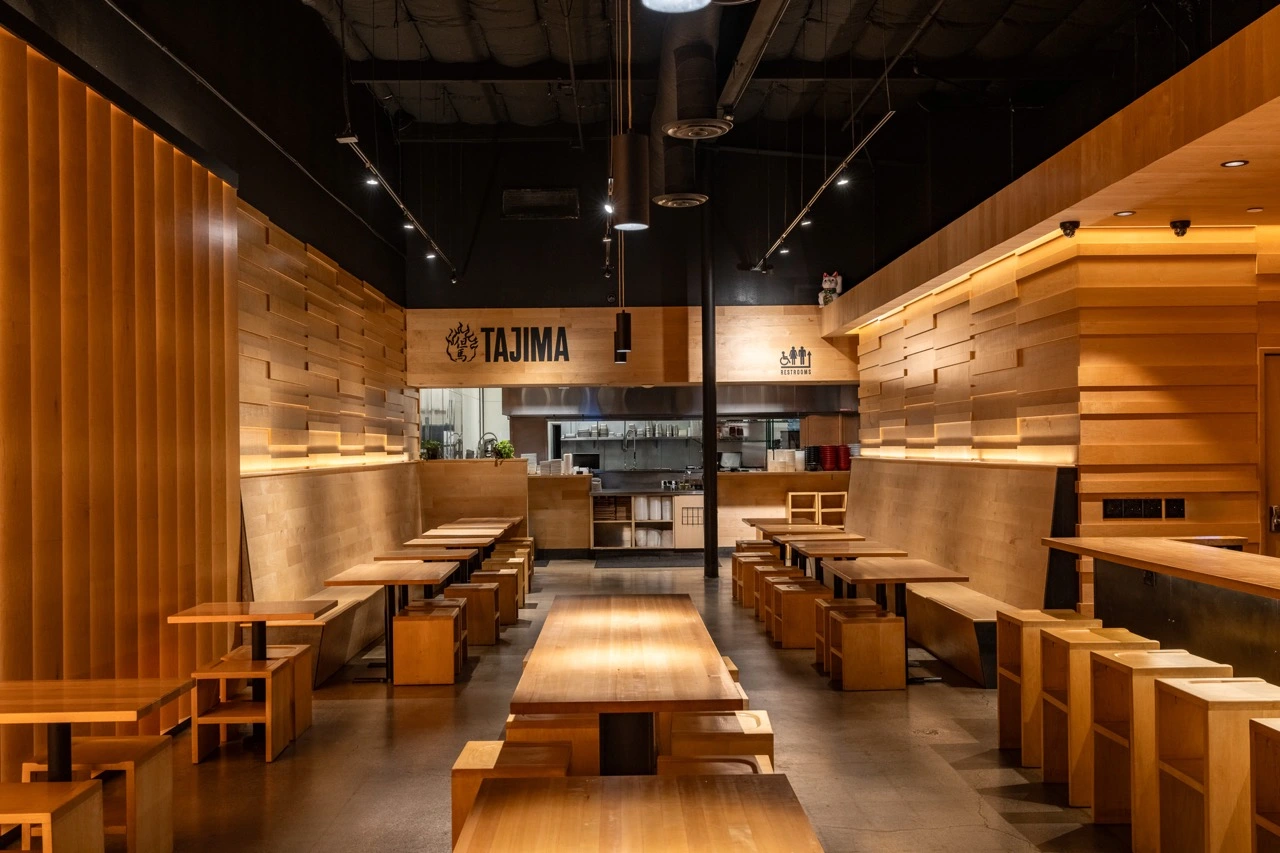 The College Heights dining room looking down the center aisle before service. Long maple communal tables run between bench seating, a screen of vertical maple slats closes the left wall, and the open kitchen sits at the far end under a black sign reading Tajima.