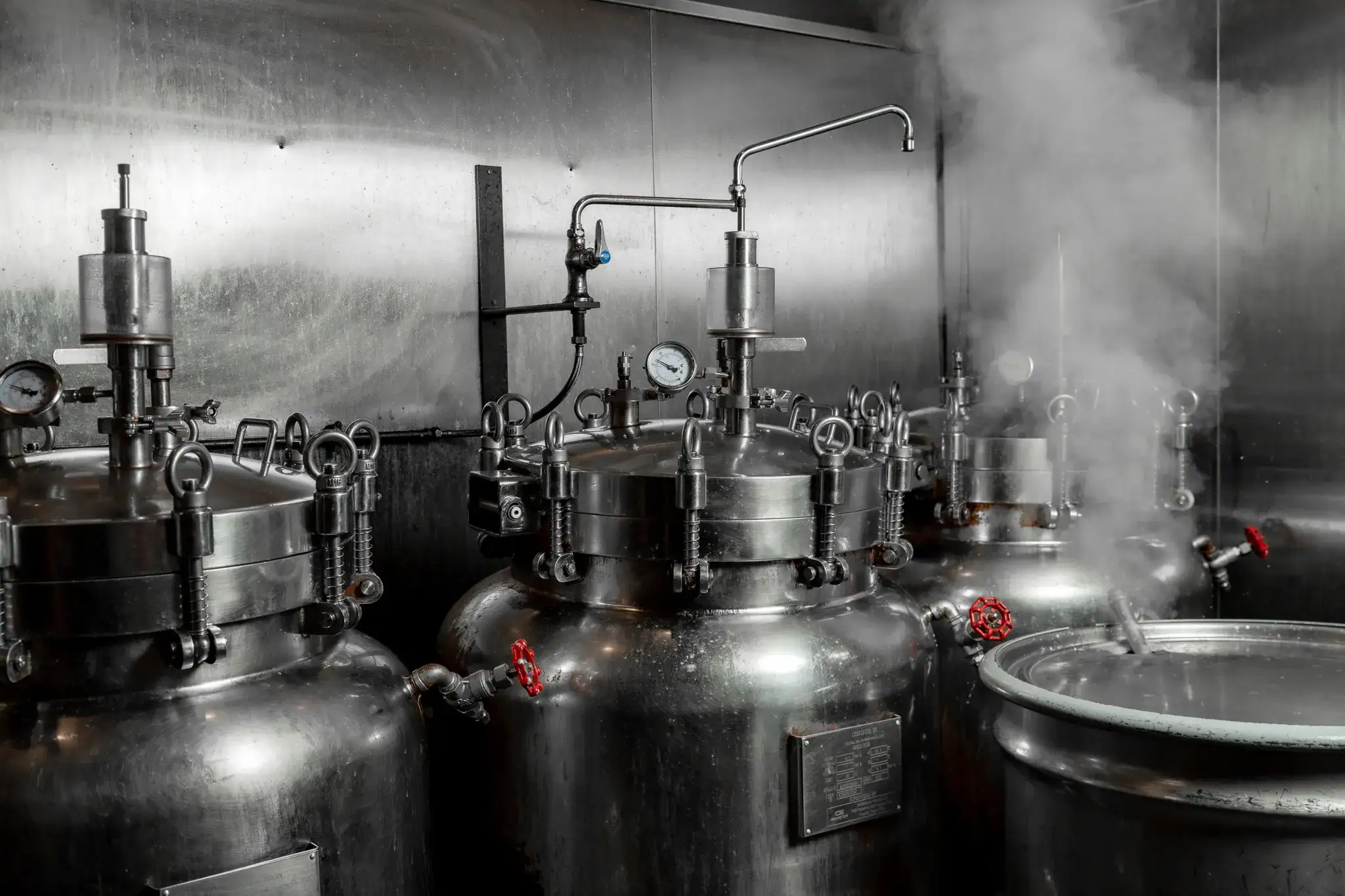 A row of stainless steel broth tanks inside the Tajima commissary, with pressure gauges, red valve handles, and steam drifting across the frame.