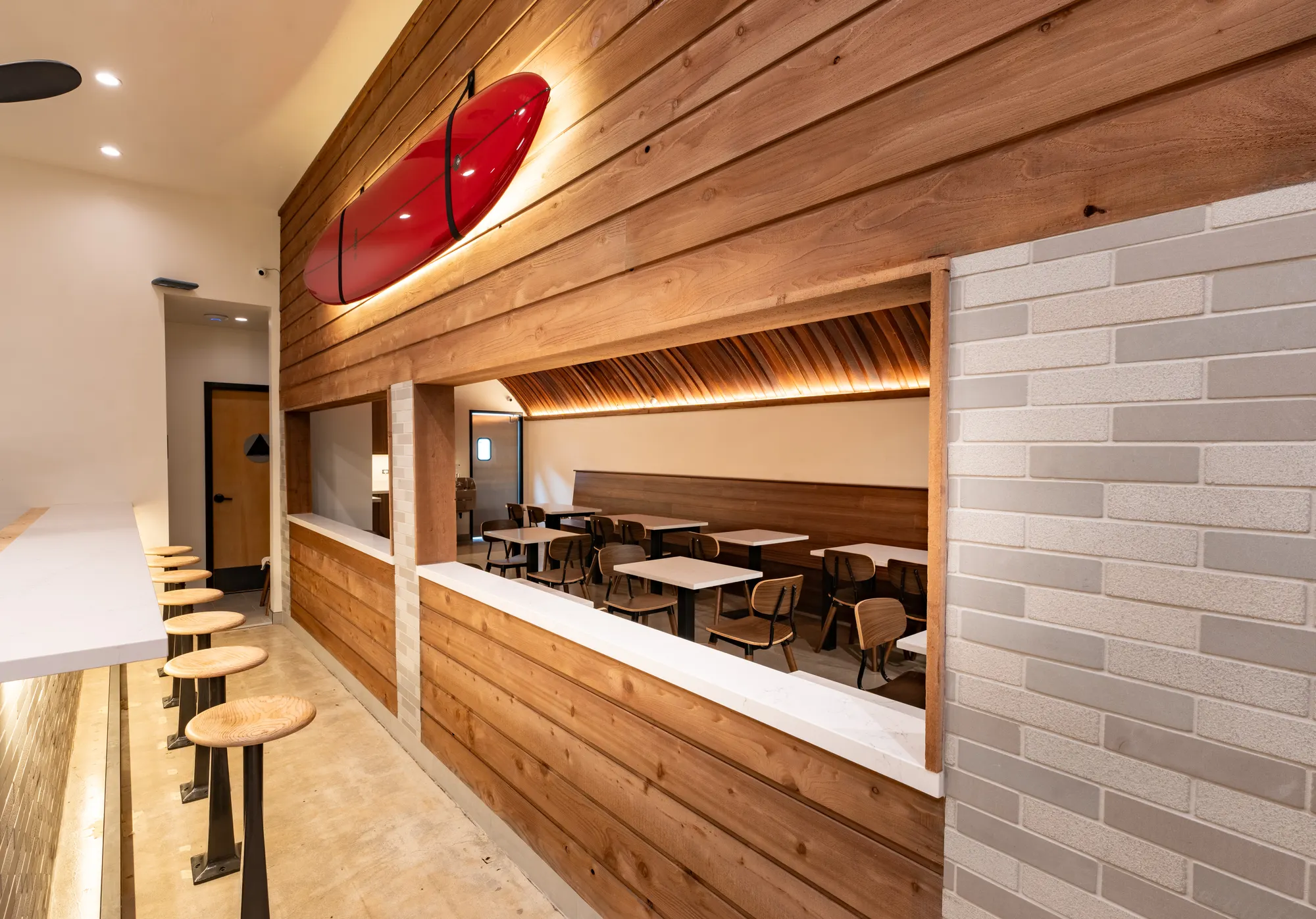 The counter at Crown Point. Wooden stools run along a white counter, with a red surfboard mounted on the cedar wall above and the dining room visible through a cut-out window.