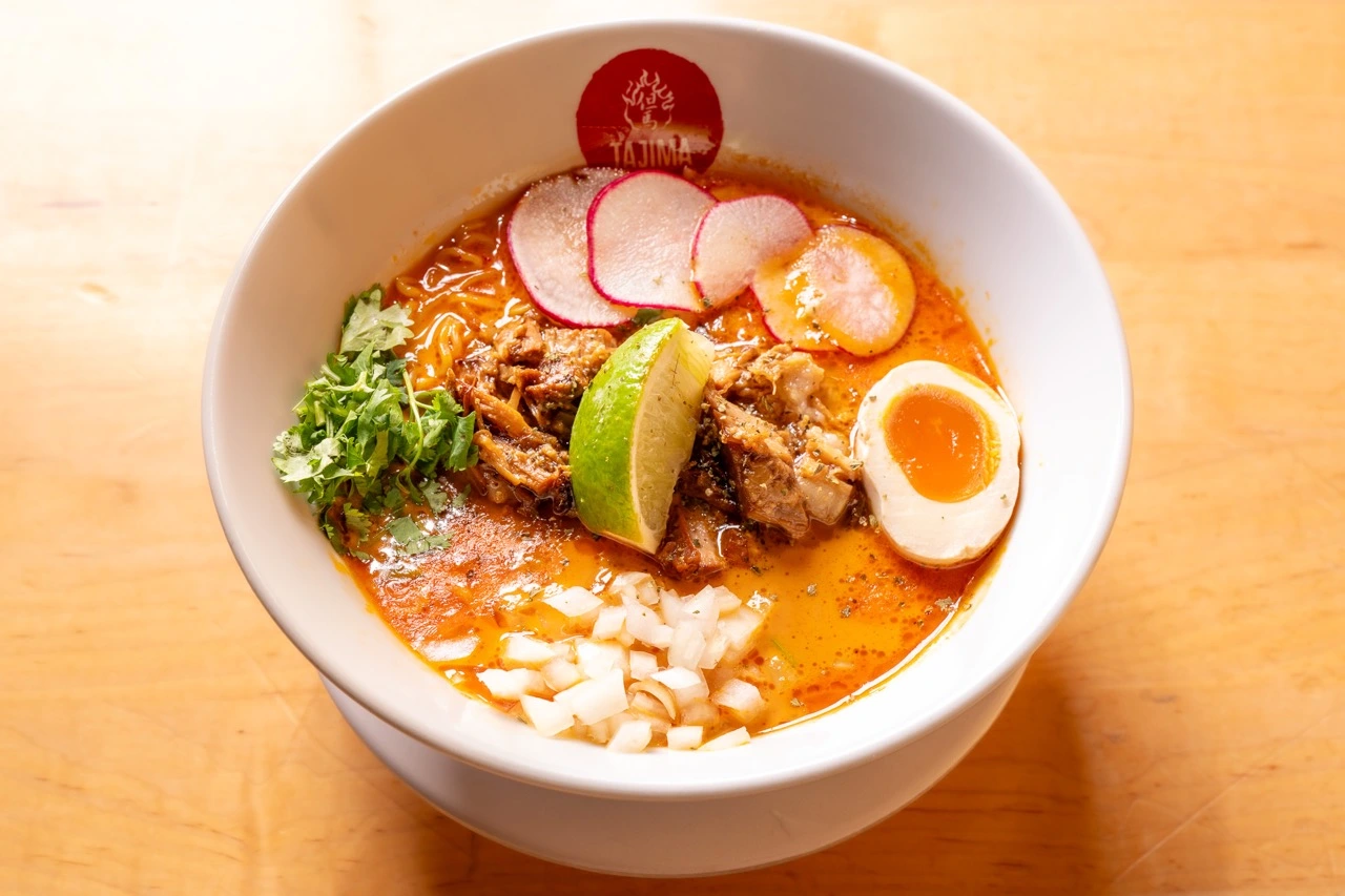 A bowl of Carnitas Ramen: noodles in a red-orange broth topped with shredded carnitas, half a soft ramen egg, sliced red radish, diced white onion, cilantro and a lime wedge, in a white bowl with a red Tajima sticker on the rim.