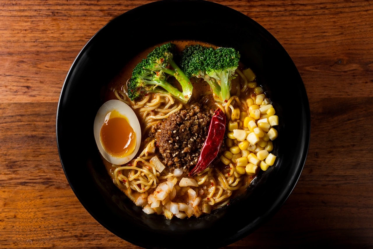 A bowl of Curry Ramen photographed from above on dark wood: curly noodles in a brown curry broth topped with seasoned ground pork, a whole dried red chili, half a soft ramen egg, two broccoli florets, sweetcorn and diced white onion, in a black bowl.
