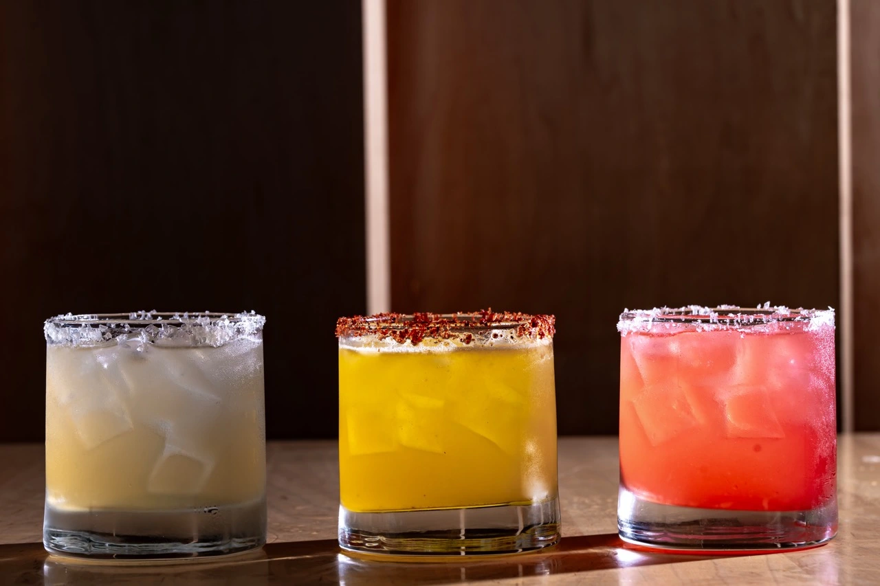 Three iced drinks in rocks glasses standing in a row on a wooden bar against a dark wood wall: a pale one with a salt rim, a yellow one with a red-flecked salt rim, and a red one with a salt rim.