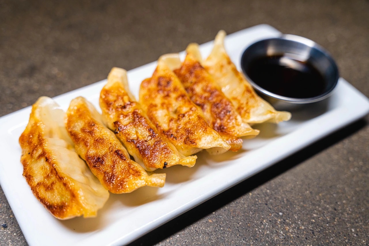 Pork Gyoza, seven pan-fried dumplings browned on one side, lined up on a white plate with a cup of dipping sauce