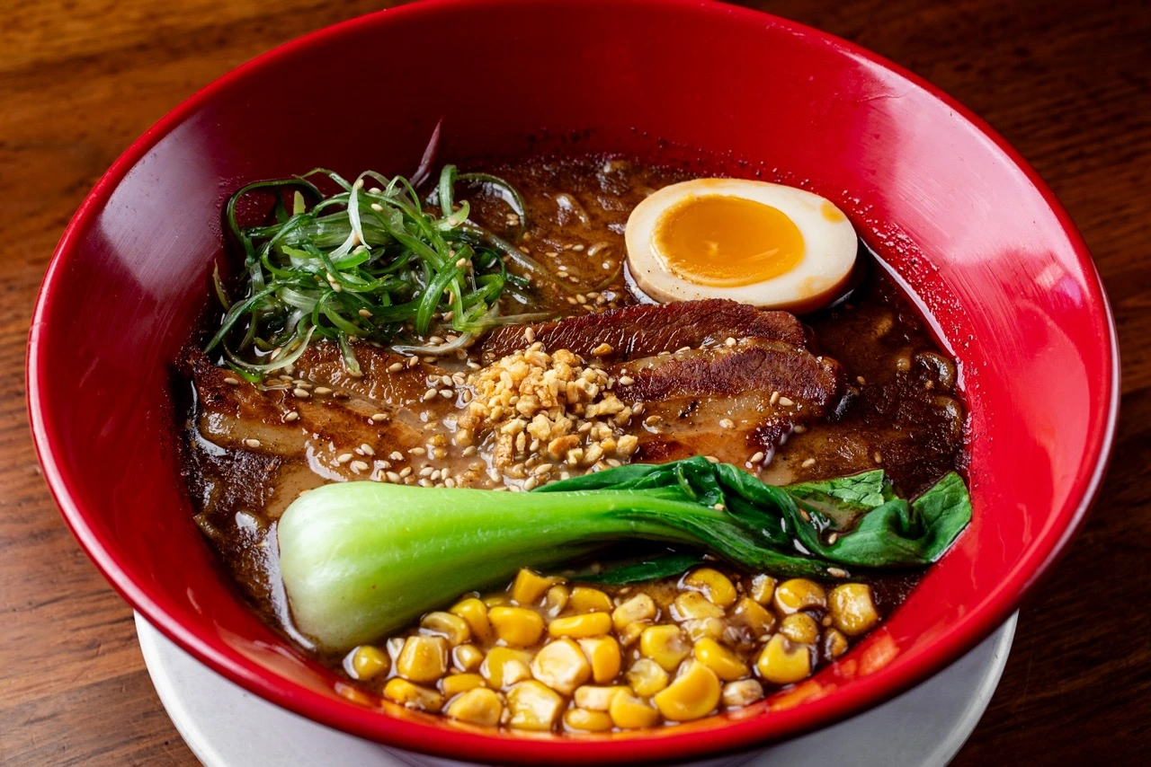 Tajima Black, two slices of pork chashu with minced garlic and sesame, half a seasoned egg, baby bok choy, sweetcorn and shredded scallion on dark black garlic broth, in a red bowl