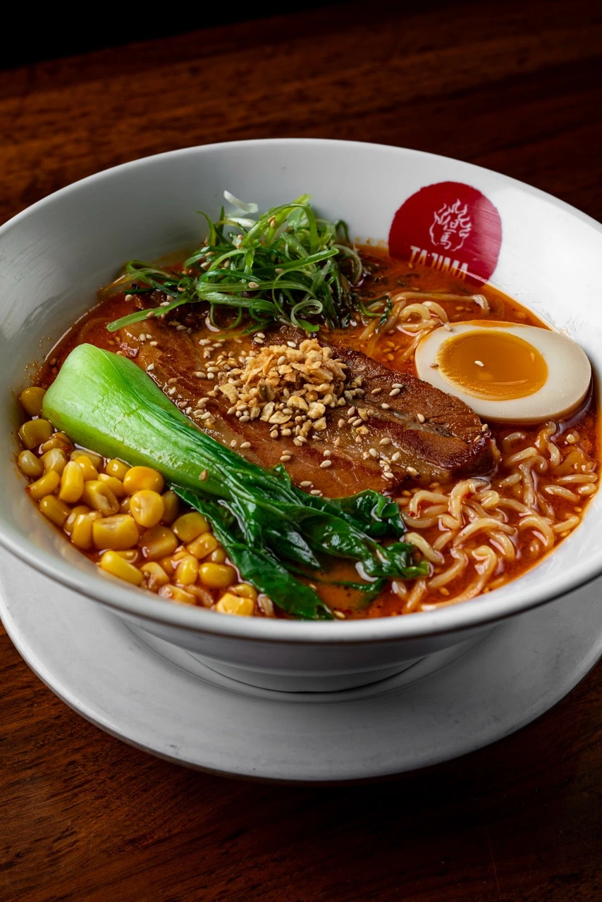 Tajima Red, a slab of pork chashu under sesame seeds and fried garlic, half a seasoned egg, baby bok choy, sweetcorn and shredded scallion on chili-red broth with ramen noodles