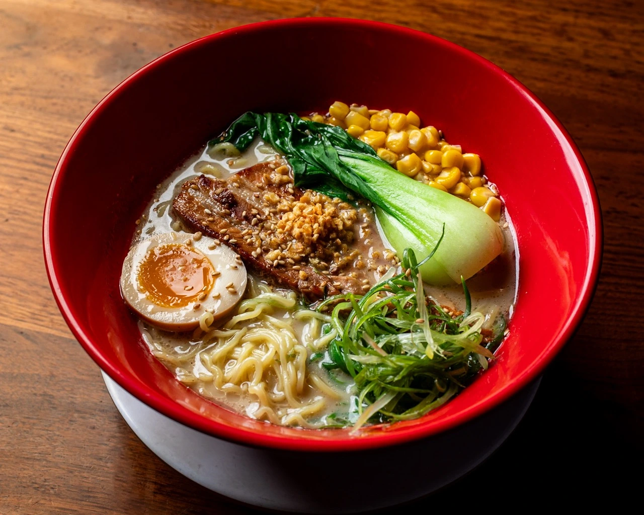 Tajima White, pork chashu under minced garlic and sesame, half a seasoned egg, baby bok choy, sweetcorn and shredded scallion on pale tonkotsu broth with wavy ramen noodles, in a red bowl
