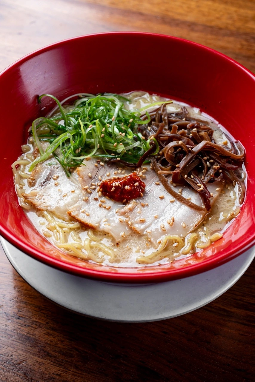 Traditional Tonkotsu, sliced pork chashu, shredded scallion, seasoned wood ear mushroom and a spoon of red chili paste on pale tonkotsu broth with ramen noodles, in a red bowl